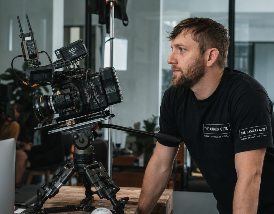 Videographer monitoring a tripod-mounted cinema camera beside a computer during an office production