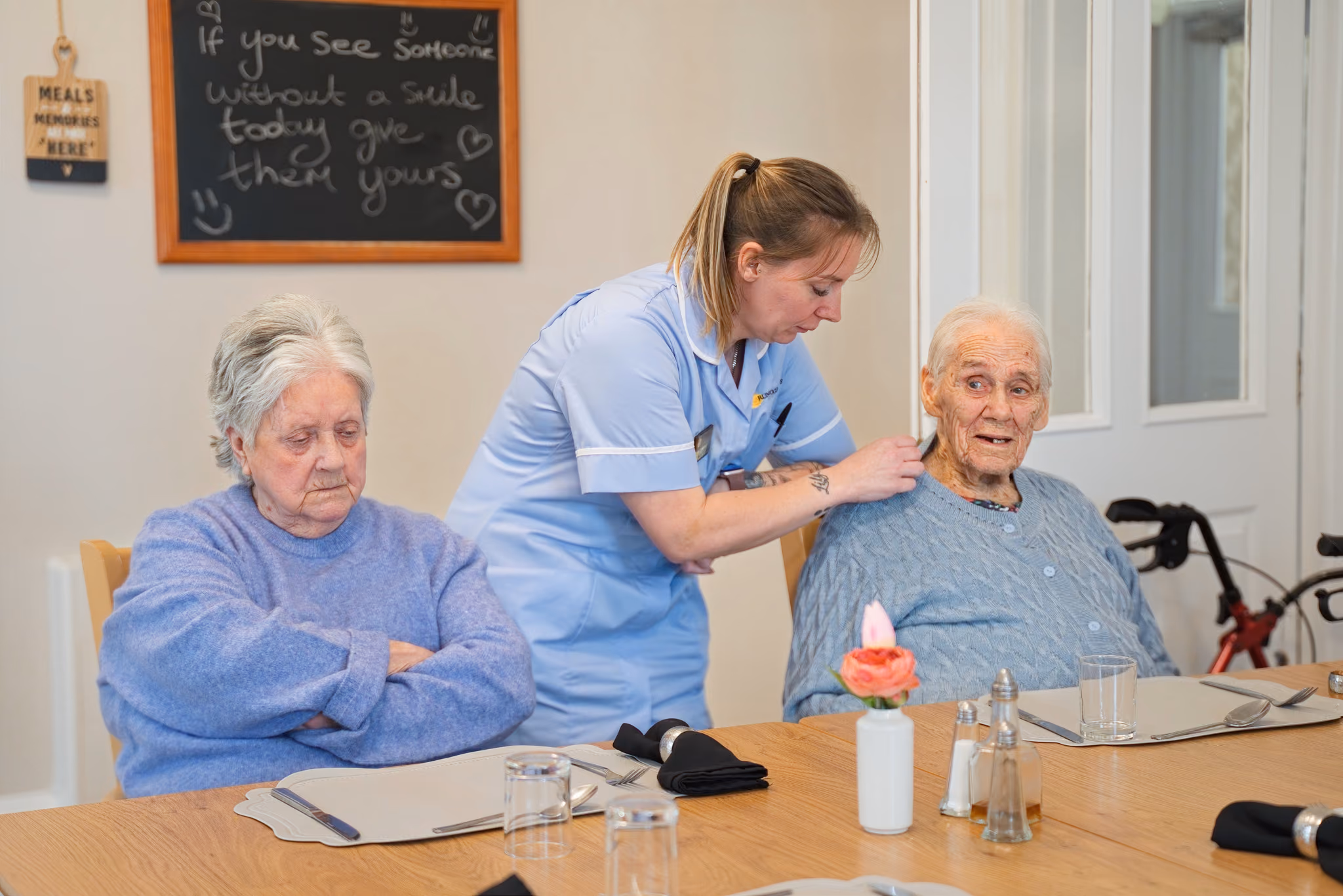 A carer leaning down to speak with a resident at the dining table