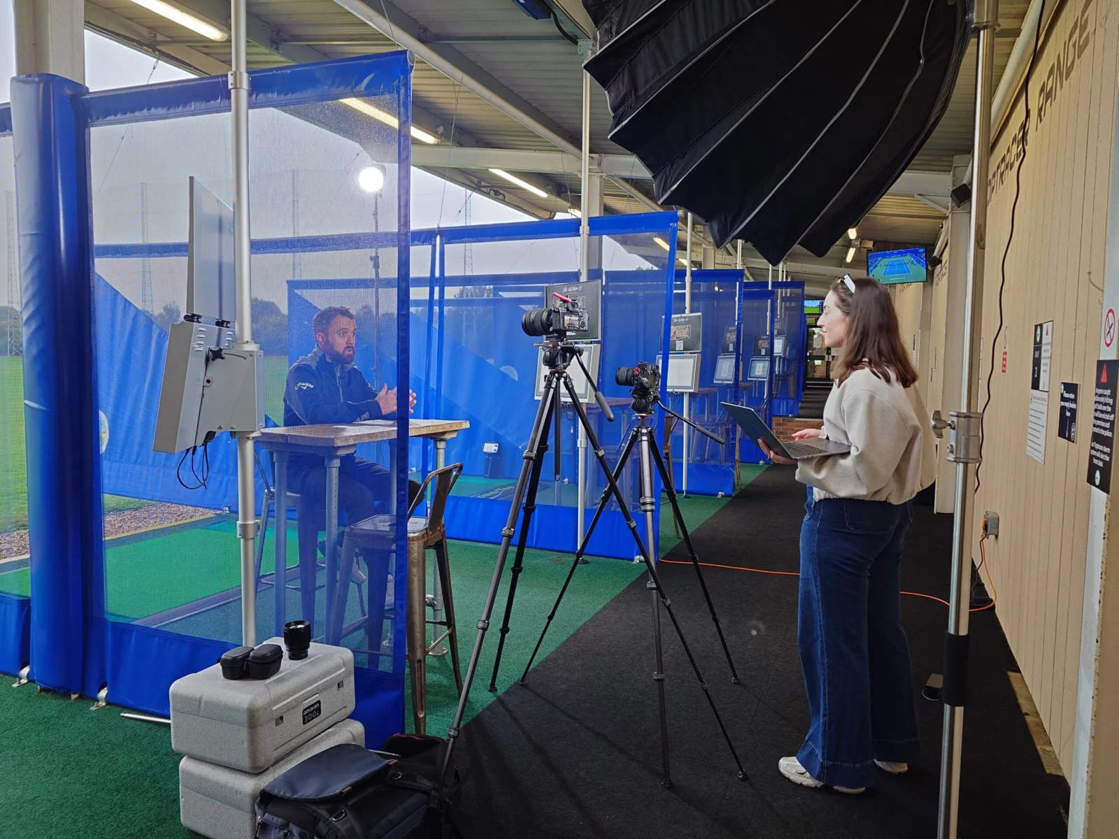 Camera operator filming a presenter at an exhibition stand, with lighting and a second camera set up
