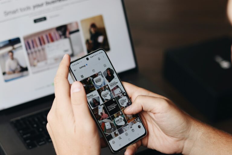 Hands holding a phone scrolling an Instagram feed next to a laptop