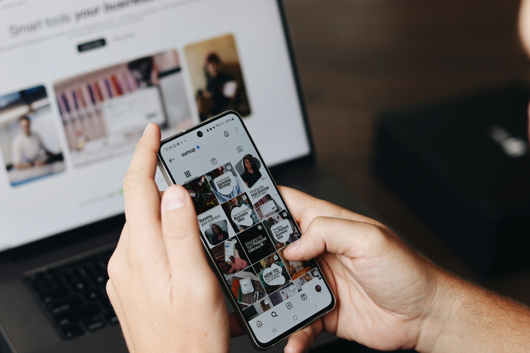 Hands holding a phone scrolling an Instagram feed next to a laptop